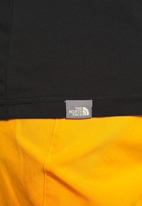 Nahaufnahme des Saums eines schwarzen Kleidungsstücks mit einem grauen "The North Face"-Logotag über einem leuchtend gelben Stoff mit sichtbaren Nähten.