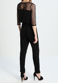 Black jumpsuit with sheer, polka-dotted sleeves and a keyhole back. Semi-fitted design, tapered legs, paired with black, ruffled high heels.