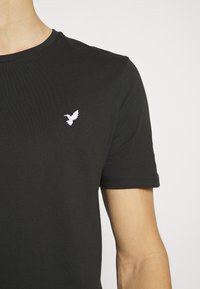 Black cotton t-shirt with a crew neck, featuring a small white embroidered bird logo on the left chest. Short sleeves, smooth texture.