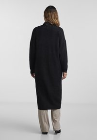 Lång svart stickad klänning med avslappnad passform, som har en rund halsringning och sänkta axlar. Slät textur, mid-calf längd, bärs över ljusbruna byxor.