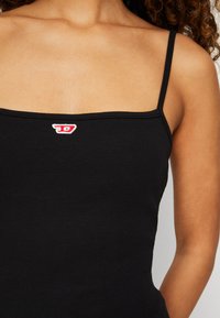 Top de alças finas, em preto, com canelados, apresentando um pequeno logotipo vermelho e branco no peito. Textura suave, design ajustado.