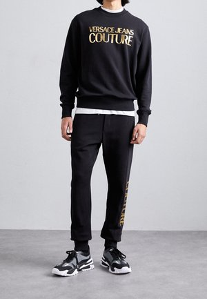 Sudadera negra con el texto dorado "VERSACE JEANS COUTURE", combinada con pantalones negros que presentan un estampado de logotipo en el lateral; llevada con zapatillas negra y blancas.