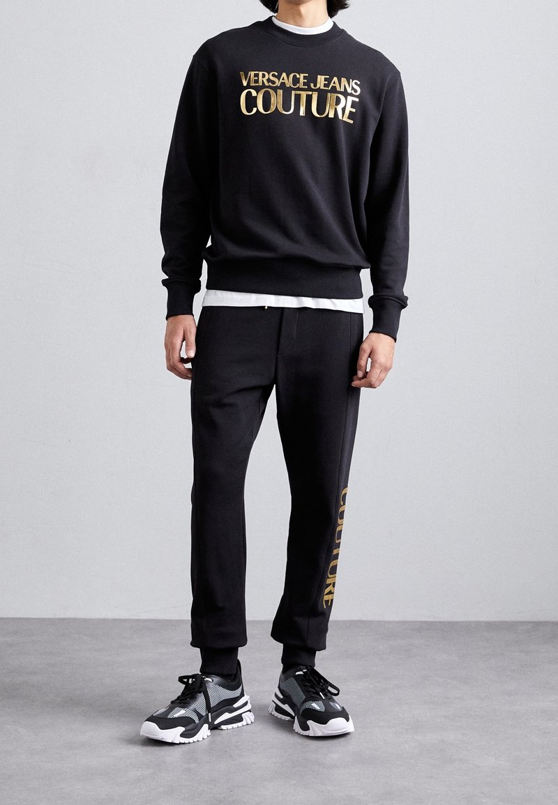 Osoba nosící černou mikinu Versace Jeans Couture a odpovídající kalhoty se zlatým nápisem, doplněné černo-bílými teniskami.