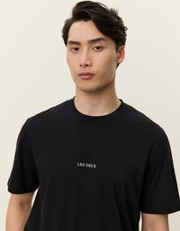Zwarte katoenen t-shirt met een ronde hals, korte mouwen en witte tekst met de boodschap "LES DEUX" gecentreerd op de borst. Gladde textuur, eenvoudig ontwerp.