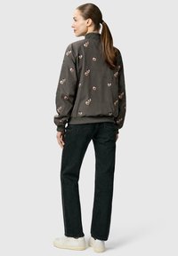 Kvinde med hestehale iført en grå bomberjakke med små blomsterprint, sorte jeans og hvide sneakers, stående med ryggen mod kameraet.