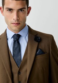 Costum maro cu o textură interesantă, cămașă albă de ocazie cu dungi fine, cravată închisă cu model și o broșă cu trandafir negru. Batistă de buzunar cu un model floral închis.