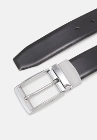 Svart läderbälte med en silverf�ärgad metallspänne som har en texturerad yta och rektangulär form. Spännet inkluderar dubbelpiggar för att fästa.