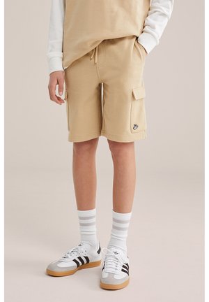 Otrok, ki nosi bež cargo kratke hlače, belo majico z dolgimi rokavi, bele nogavice s črtami in bele superge s črnimi črtami.