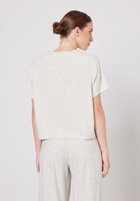 Juuksed kuklas, seljaga vaatajate poole, seljas helehall lühikeste varrukatega särk ja sobivad lahtised püksid, tavalise tausta ees.