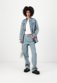 Svetlo modra jeans jakna z dvema žepoma na prsih, v kombinaciji z usklajenimi visokimi hlačami in črnimi platform čevlji, drži bež torbico.