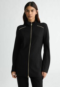 Črna zip-up jakna z visokim ovratnikom, ki ima zlate zadrge in stranske žepe. Gladek material in kroj, ki se prilega.