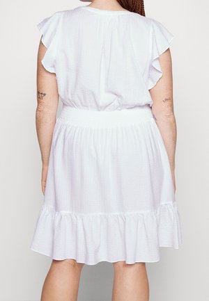 Woman wearing a white textured dress with flutter sleeves and ruffled hem, shown from the back against a plain background.