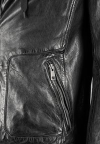 Zwarte leren jas met een textuurafwerking, een voorkantzak en een zijzak met rits en metalen hardware. Versterkte stiksel-details.