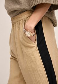 Beige bukser med en sort sideliste, blød tekstur og elastisk talje, som holder et hvidt og gyldent ur i en sidelomme.