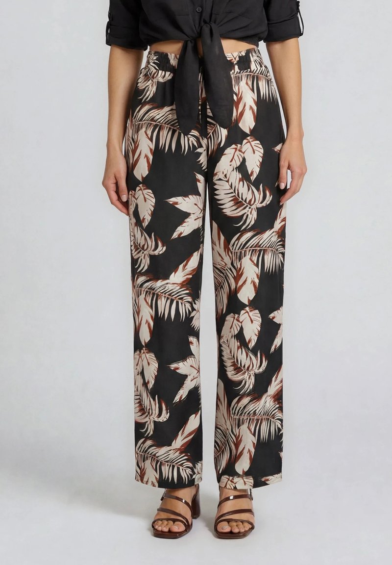 Femme portant un pantalon noir à jambes larges avec un imprimé feuilles tropicales beige et marron et des sandales à talons marron à brides.