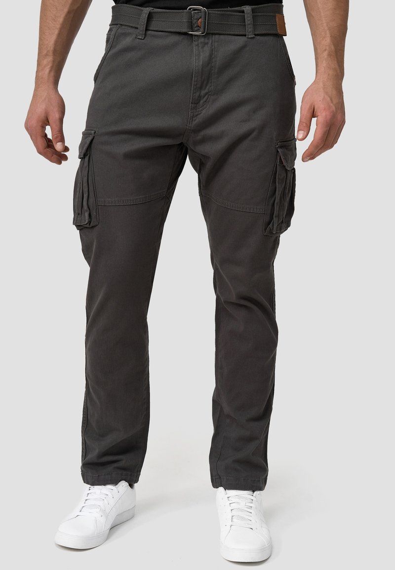 Homme portant un pantalon cargo gris foncé avec poches latérales, une ceinture noire tissée et des baskets blanches, debout devant un fond neutre.