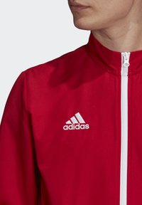 Rød atletisk zip-up jakke lavet af glat stof. Har en hvid lynlås og et broderet hvidt Adidas-logo på venstre side.