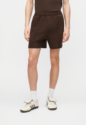 Personne portant un short marron foncé, un haut sans manches marron assorti, des chaussettes blanches et des baskets blanches avec des rayures noires.