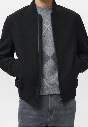 Mand iført sort lynlåstrøje over grå argyle sweater og falmede grå jeans, hænderne i jakke lommerne, står mod ensfarvet baggrund.