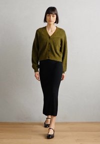 Gilet tricoté vert olive avec un col en V et une fermeture à boutons, associé à une jupe midi noire côtelée et des chaussures Mary Jane noires.
