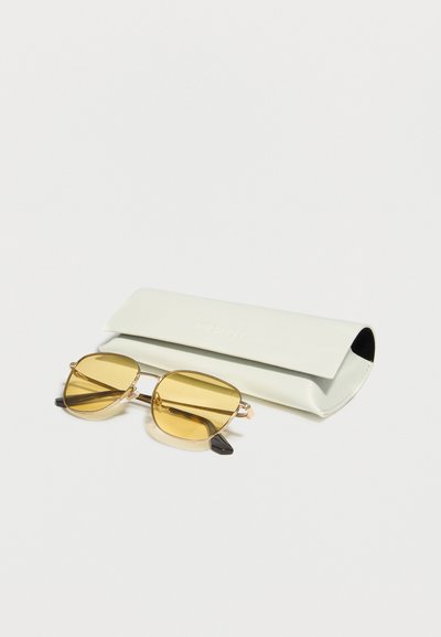 Γυαλιά ηλίου με χρυσό σκελετό και κίτρινους φακούς που τοποθετούνται μπροστά σε λευκή θήκη γυαλιών από δέρμα, πάνω σε λευκό φόντο.