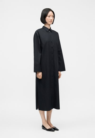 Γυναίκα με κοντά μαύρα μαλλιά που φοράει μακρυμάνικο midi φόρεμα με κουμπιά μαύρου χρώματος και μαύρες μυτερές μπαλαρίνες, στέκεται μπροστά σε απλό λευκό φόντο.