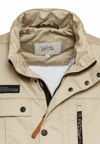 Beżowa kurtka z wysokim kołnierzem, czarnym zamkiem błyskawicznym i zatrzaskami. Posiada dwie naszywane kieszenie oraz metkę z logo wewnątrz. Teksturowana tkanina.