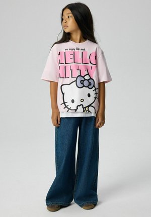 Dziewczyna z długimi czarnymi włosami, ubrana w różowy t-shirt Hello Kitty i szerokie niebieskie dżinsy, stojąca i patrząca w lewo.
