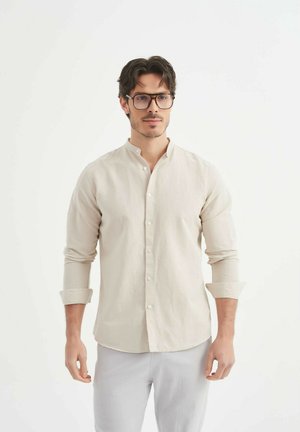 Chemise beige clair à boutons avec un col mandarin, longues manches retroussées aux poignets, en tissu texturé, portée sur un pantalon gris clair.