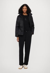 Black padded jacket with a hood, featuring a subtly patterned fabric. Worn over a black T-shirt and black sweatpants. Beige shoes with light accents.