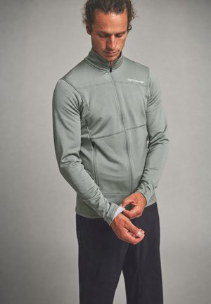 Man adjusting cuff of light grey zip-up long-sleeve jacket with Ortovox logo, wearing black pants, standing against plain grey background.