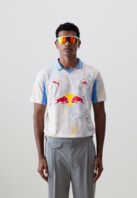 Bijela i svijetloplava majica s cvjetnim uzorkom i Red Bull logotipom. Uparena sivoj hlačama. Model nosi šarene sunčane naočale.