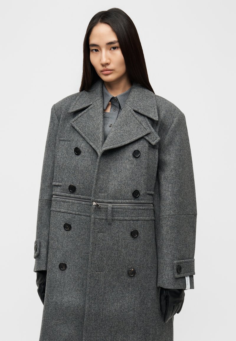Manteau en laine grise à double boutonnage avec grands revers crantés, six boutons noirs, poches latérales et détail zippé à la taille. Gants noirs portés.