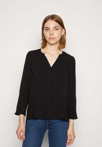 Maglietta nera a maniche lunghe con scollatura a V e leggera texture. Presenta una vestibilità rilassata e spacchi laterali. Abbinata a jeans blu.