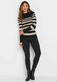 Gestreifter Strickpullover in Schwarz und Beige, mit hohem Kragen, langen Ärmeln und Knopfdetails an den Bündchen. Kombiniert mit schwarzen Hosen und Stiefeln.