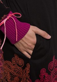 Vêtement noir avec des motifs orange ornés à l'ourlet. Manchon violet côtelé avec un accent en ruban rose. Main dans la poche latérale.