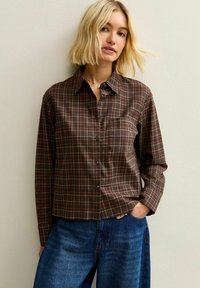 Camicia a quadri marrone con colletto, taschino anteriore e maniche lunghe, abbinata a jeans in denim blu.