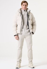 Pufferjack met een lichte beige kleur en een capuchon, met een gequilt ontwerp en zwarte accenten, gedragen over een grijze kabeltrui en lichtbeige broeken.