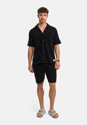 Schwarzes, kurzärmeliges Hemd mit Knopfleiste und Kragen, kombiniert mit passenden Shorts. Das Material scheint strukturiert zu sein. Getragen mit grauen Sandalen.