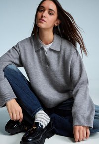 Grauer Strickpullover mit kragenartigem Halsauschnitt, umgeschlagenen Bündchen, kombiniert mit blauen Jeans und glänzenden schwarzen Slippern sowie weißen gestreiften Socken.