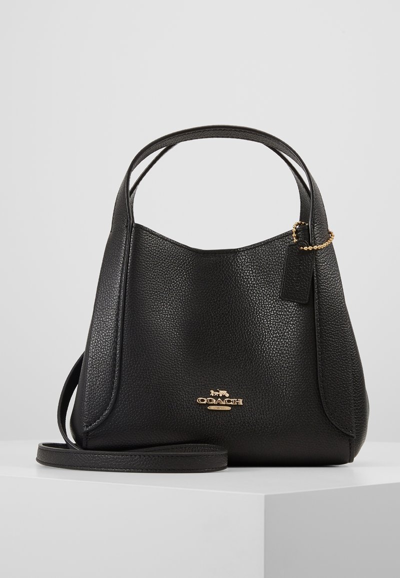 Sac à main Coach en cuir noir avec poignées courtes, bandoulière amovible et logo ainsi qu’étiquette dorés sur une surface blanche.