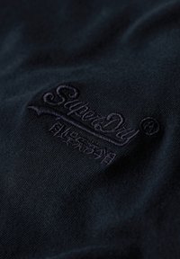 Sort stof med en glat tekstur, der har en struktureret mørk brodering af "Superdry"-logoet i hjørnet.