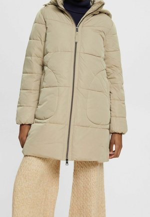 Personne portant un manteau matelassé à capuche beige avec une fermeture éclair à l'avant et des poches arrondies, assorti à un pantalon large en maille crème.