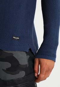 Maglione a maglia testurizzata blu navy con etichetta "Only & Sons" abbinato a pantaloni mimetici neri e grigi, mostrando una mano rilassata al lato.