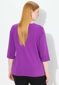 Lila T-Shirt mit Dreiviertelärmeln, glatter Textur, Rundhalsausschnitt und lockerem Schnitt, kombiniert mit schwarzen Hosen.