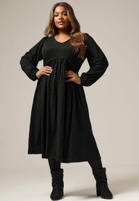 Vestido negro texturizado de manga larga con escote en V, cintura imperio y falda fluida. Combinado con botas negras hasta el tobillo, creando un look en capas.