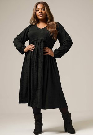 Czarna, fakturowana sukienka z długim rękawem, dekoltem w kształcie litery V, empire waist i zwiewną spódnicą. Połączona z czarnymi botkami za kostkę, tworząc warstwowy wygląd.