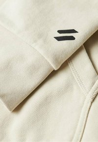 Beige sweatshirt fabric with visible texture and stitching. Features a black logo on the collar. Smooth, thick fabric.