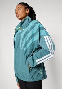 Zelena jakna od windbreakera s bijelim prugama, visokim ovratnikom, zatvaranjem na zip, i bočnim džepovima. Ima laganu, strukturiranu tkaninu i detalje s logotipom.