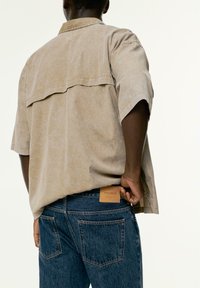 Chemise beige à manches courtes avec un détail de passepoil au dos, portée sur un jean en denim bleu foncé avec une étiquette en cuir et des poches arrière.